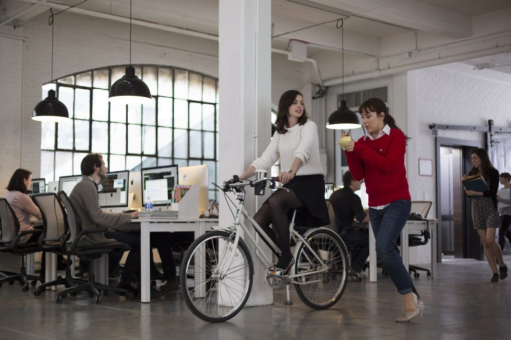 Imagem promocional do filme "Um Senhor Estagiário", disponível na HBO Max. Nela, vemos duas mulheres, sendo uma delas Jules Ostin, a protagonista, em um escritório. Jules está sentada em uma bicicleta, algo atípico para o ambiente corporativo, e a outra mulher, que parece ser sua estagiária, está correndo atrás dela e fazendo anotações.