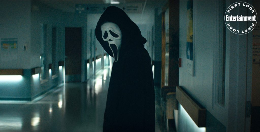 O assassino ghostface está no meio de um corredor de hospital vazio. Ele está de lado, olhando para o chão.