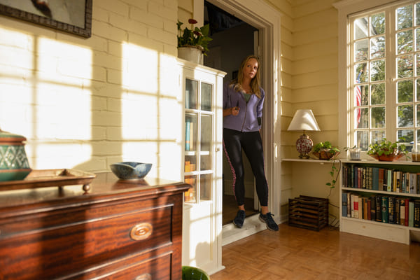 Uma sala de estar bem ampla e iluminada pela luz solar, há uma mulher loira na porta adentrando à sala com certo receio