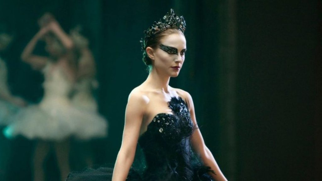 Na imagem, Natalie Portman interpreta o Cisne negro no filme de mesmo nome. Ela está vestida como uma bailarina, com um vestido e maquiagem pretas. No fundo desfocado, há outras bailarinas vestidas na cor branca.