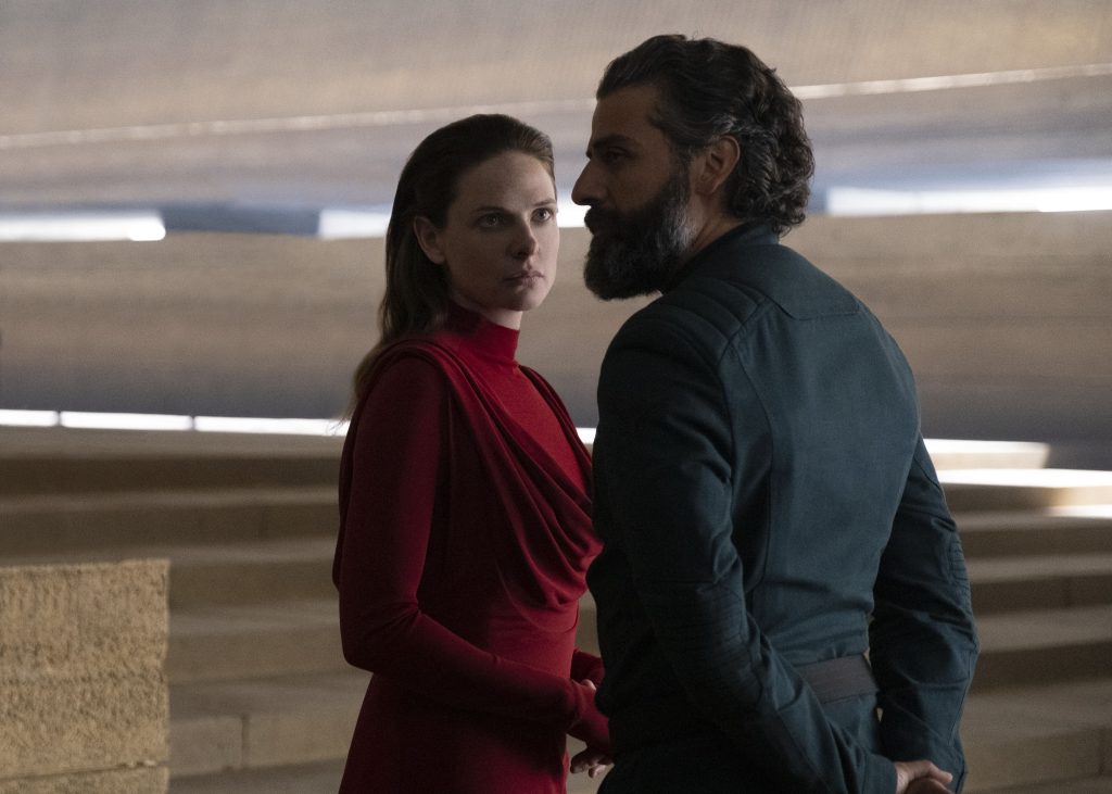 Imagem promocional de "Duna" (2021), onde vemos Rebeca Ferguson como Lady Jessica, à esquerda e de vermelho, encarando Oscar Isaac, que interpreta o duque Leto Atreides, à direita e com as costas viradas para a câmera.