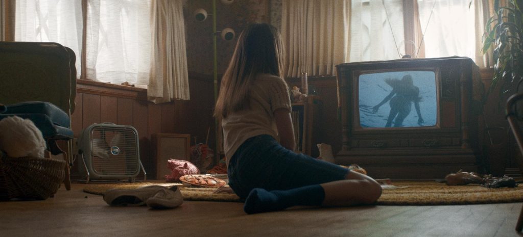 Mulher sentada sobre um tapete no chão de uma sala, ela está diante de uma televisão