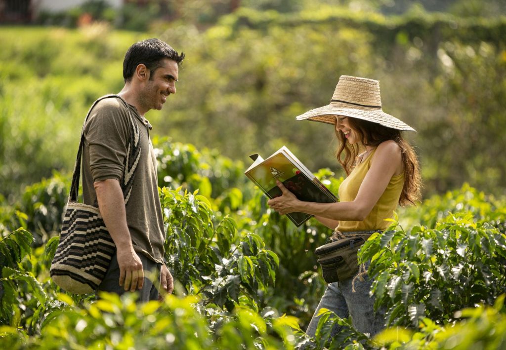 Um casal  em meio a um campo, o homem está sorrindo com uma bolsa do lado e a mulher está com um livro em mão lendo