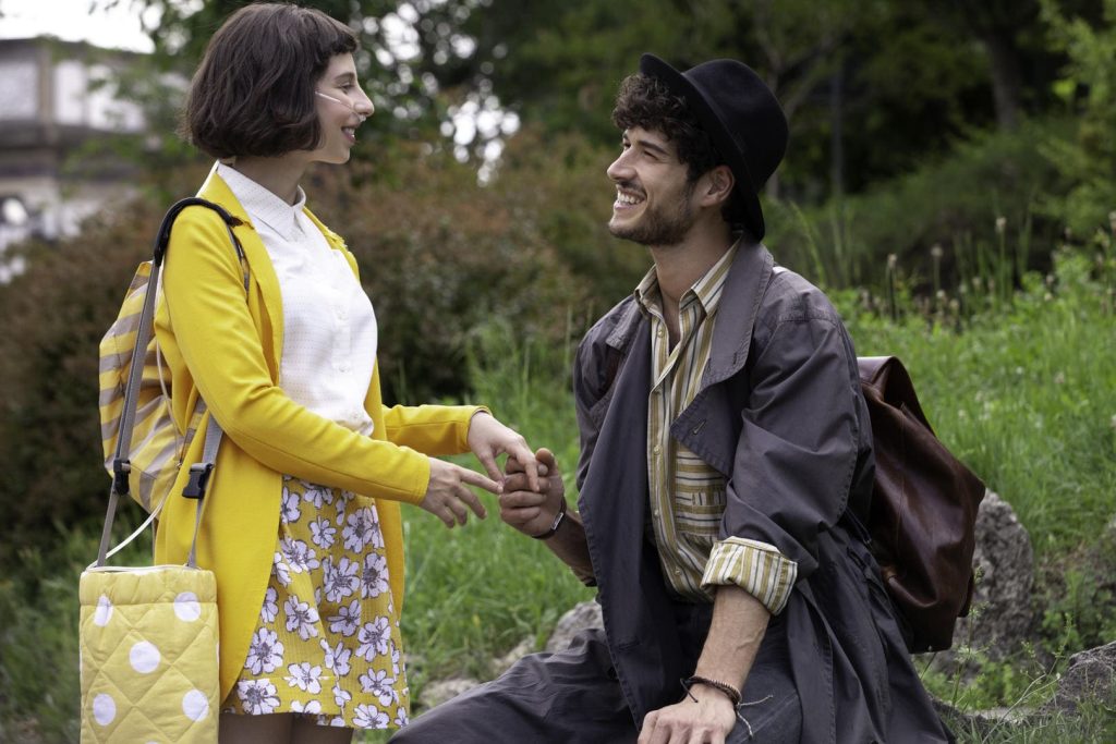 Um casal: a mulher está com roupas amarelas e uma bolsa ao lado, olhando sorridente para homem ajoelhado à sua frente, segurando suas mãos. Ele está com roupas escuras e chapéu e mochila