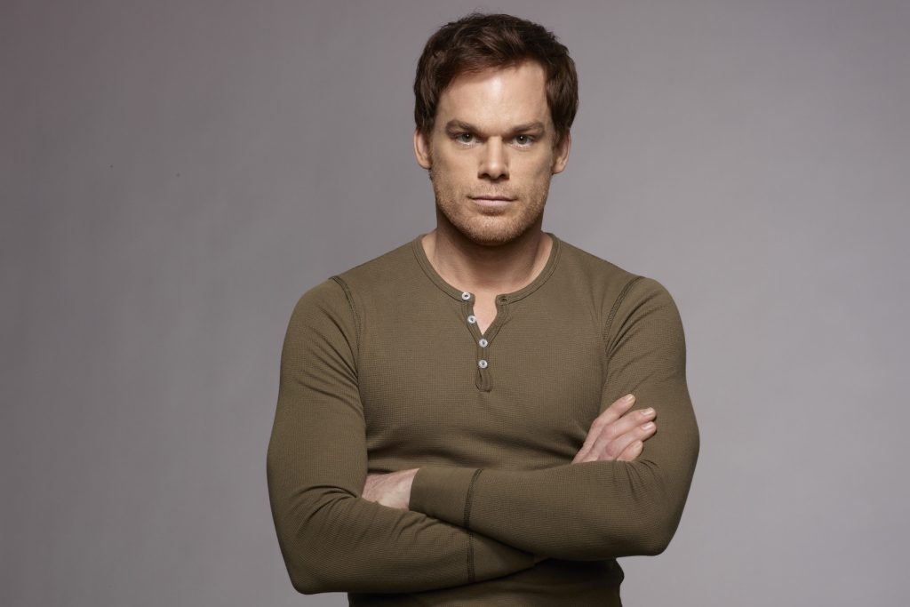 Imagem promocional da série "Dexter", onde vemos o protagonista, homônimo, usando uma camisa marrom, de braços cruzados de frente a uma parede branca, encarando a câmera