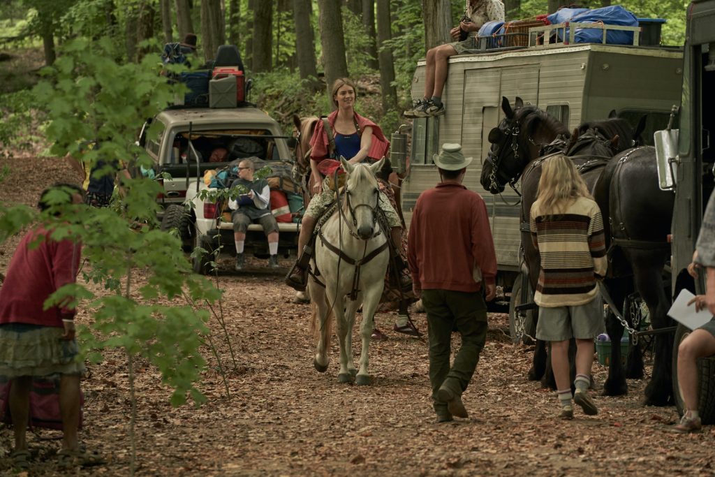 Imagem promocional da série "Station Eleven". Nela, vemos uma moça andando de cavalo em uma mata, em meio a uma comunidade, com carros, pessoas e outros cavalos em sua volta.