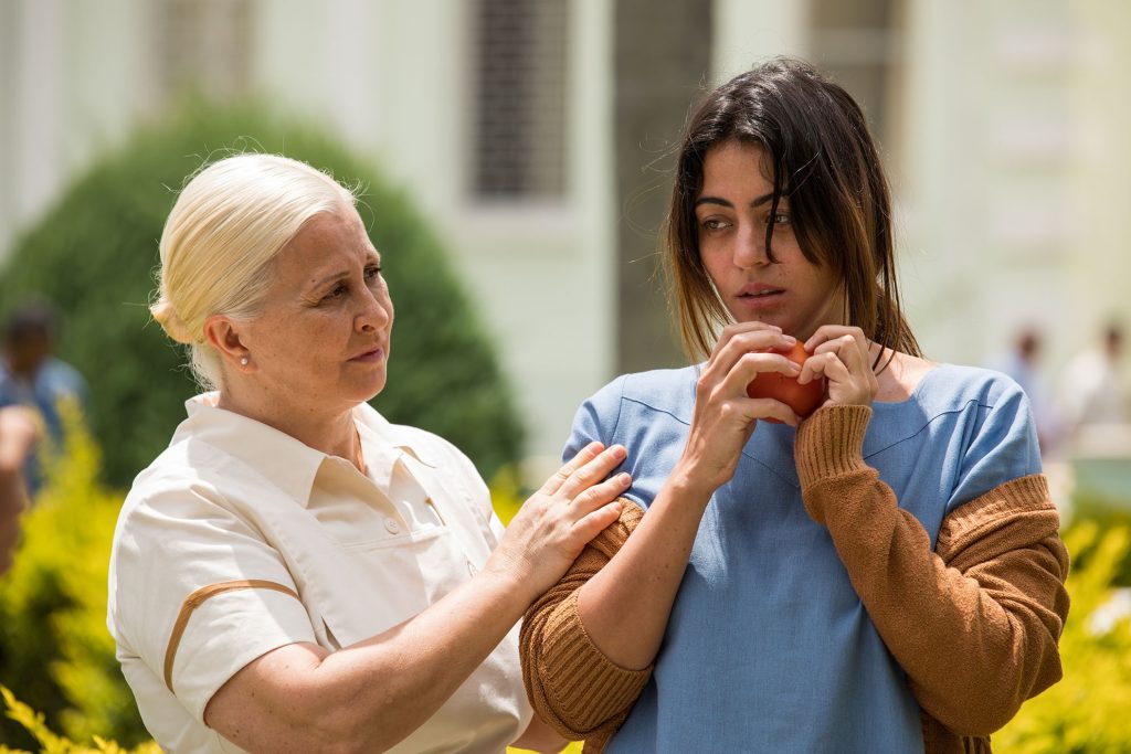 Imagem promocional da série "Insânia", do Star+. Nela, vemos uma senhora, de cabelos grisalhos, que parece guiar uma moça, com roupa de paciente hospitalar, que está com aparência confusa, e segura uma maçã em suas duas mãos