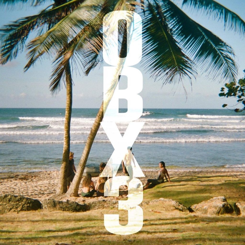 Banner de divulgação da terceira temporada de Outer Banks. Os personagens estão sentados próximo ao mar em um gramado da ilha. No meio da tela e na vertical, está escrito "OBX3", a abreviação para Outer Banks 3.
