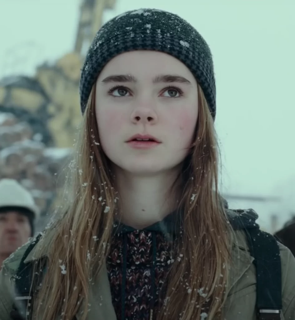 A personagem Maisie, na faixa dos 15 anos, em um lugar frio e nevando com pessoas ao redor.