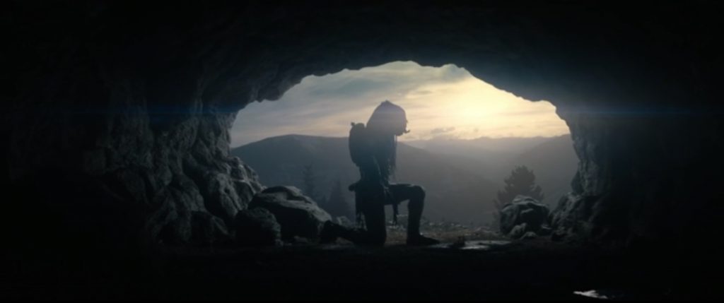 A cena mostra o Yautja ajoelhado de perfil em sua caverna tendo o horizonte ao fundo.