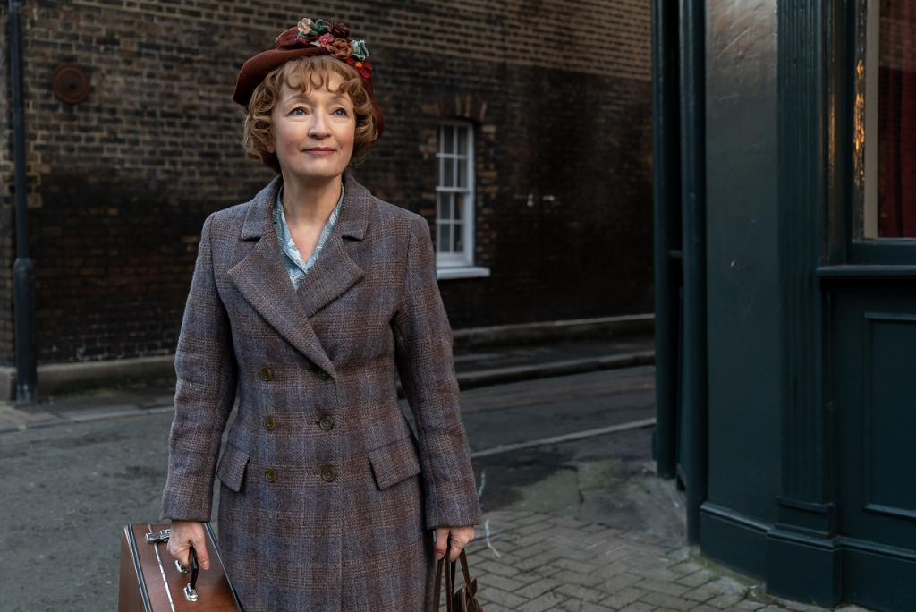 Imagem promocional de Sra. Harris Vai a Paris. Nela, cemos Lesley Manville, caracterizada como a personagem-título, vestindo um sobretudo cinza e carregando malas nas duas mãos, na Londres dos anos 1950