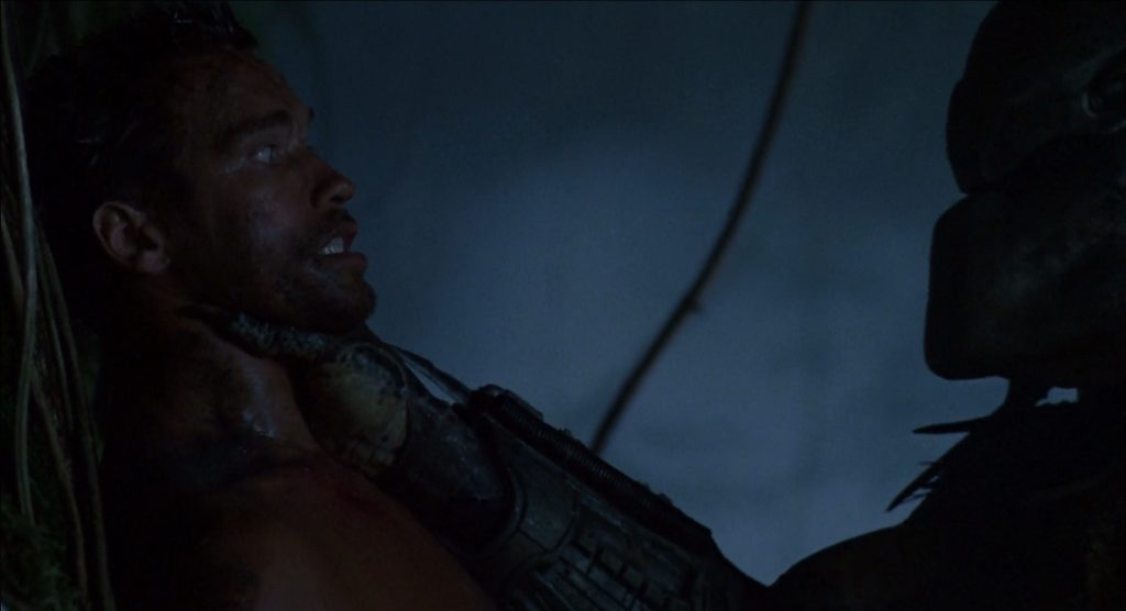 O yautja segura o Major Alan Dutch Schaeffer pelo pescoço e o encara, no primeiro filme O Predador, de 1987.