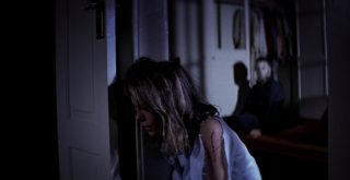 Mulher encostada na batente de uma porta enquanto um homem se levanta atrás dela